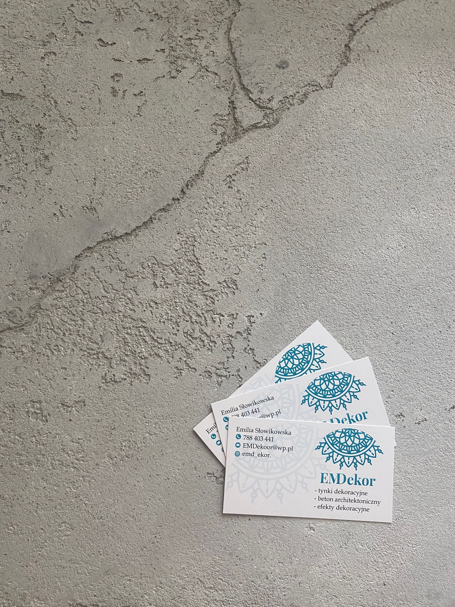 Dekoracyjny beton architektoniczny w tle z wizytówkami firmy EMDekor oferującej tynki i efekty dekoracyjne. Minimalistyczny design i stonowana kolorystyka.