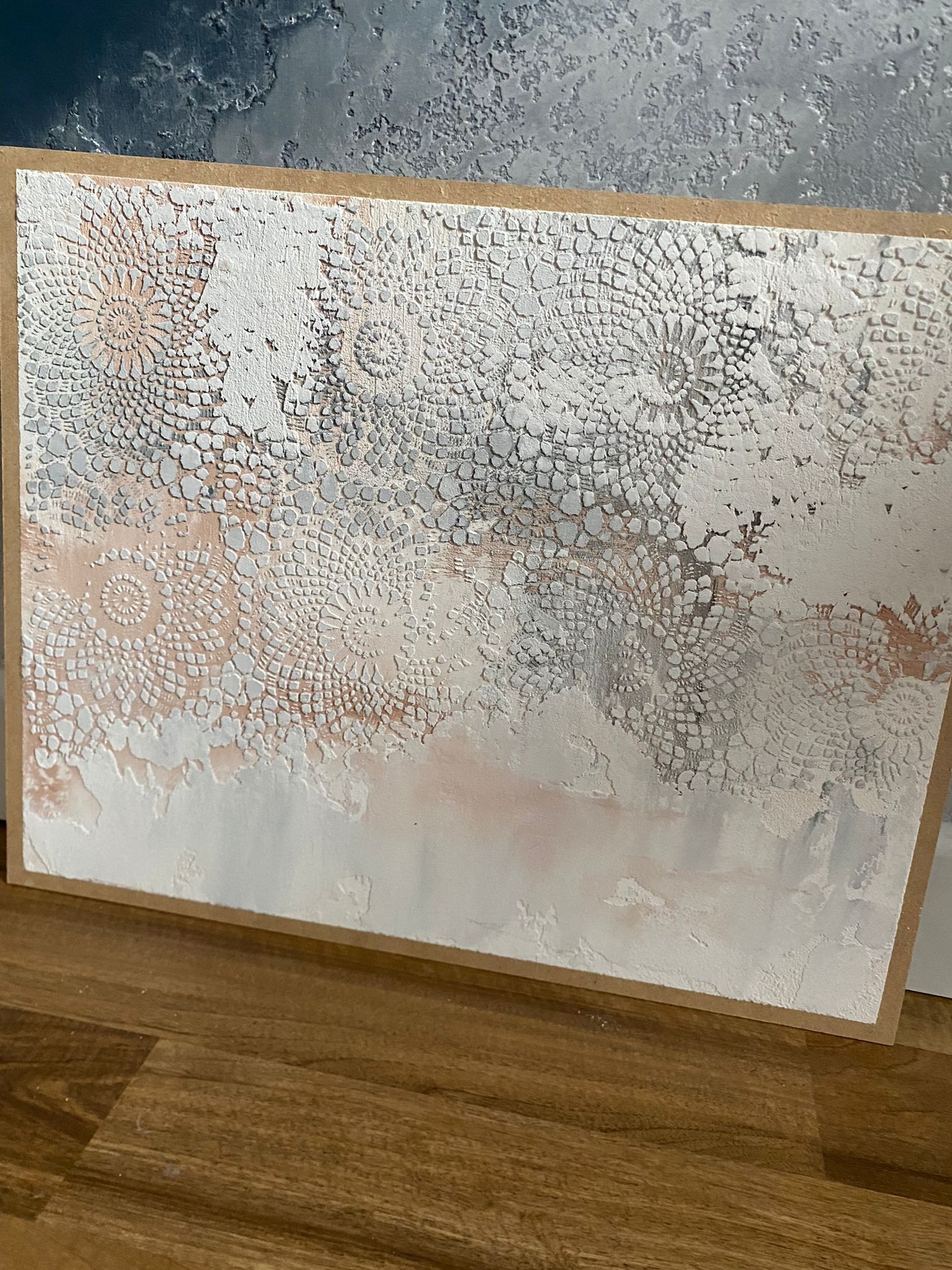 Dekoracyjna faktura ściany z motywem koronki w odcieniach bieli, szarości i różu, na drewnianym podłożu. Efektowna technika tynkarska.