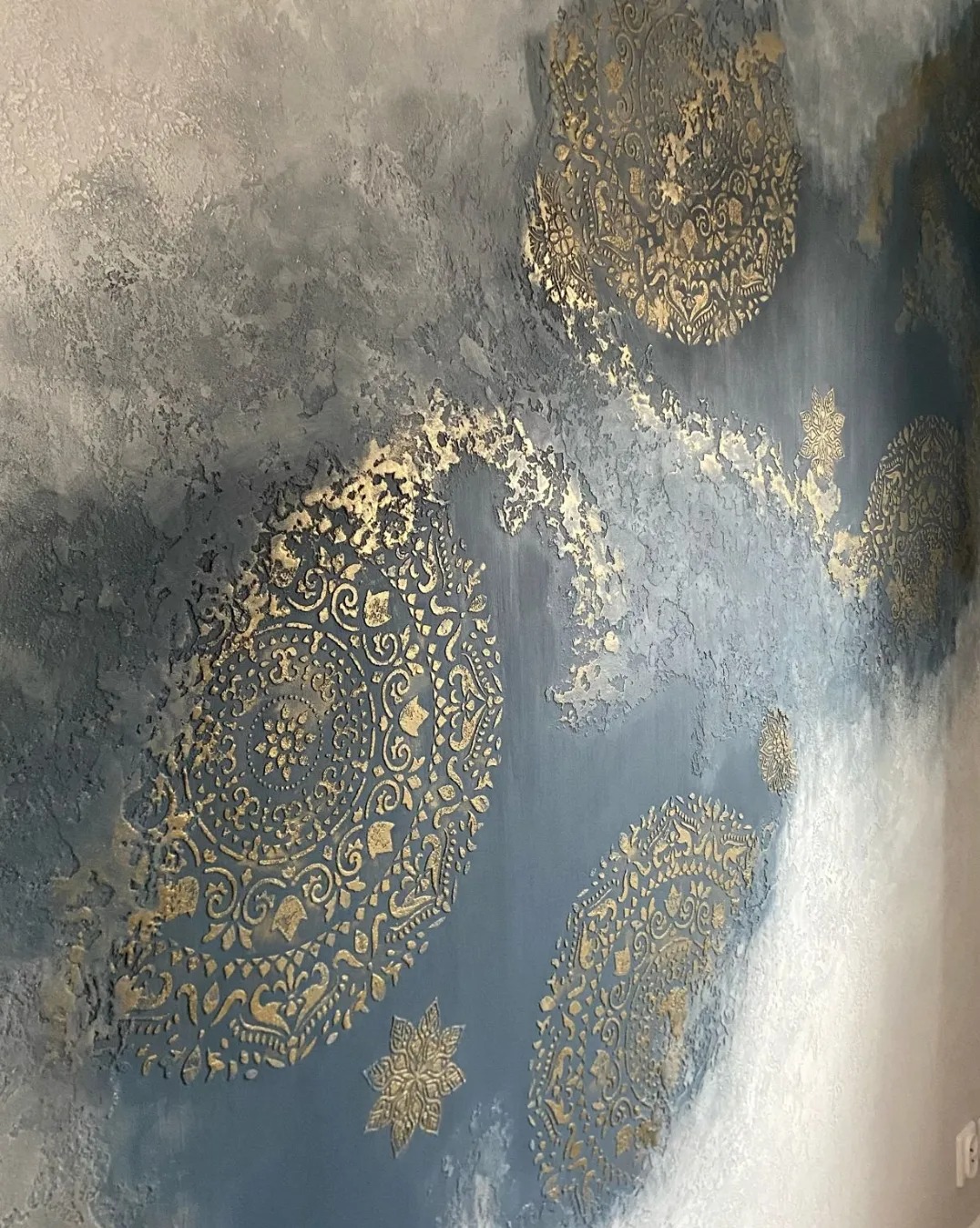 Dekoracyjna ściana z efektem ombre w odcieniach szarości i błękitu, zdobiona złotymi, wypukłymi ornamentami w stylu mandali i gwiazd. Unikalna faktura i wzór.