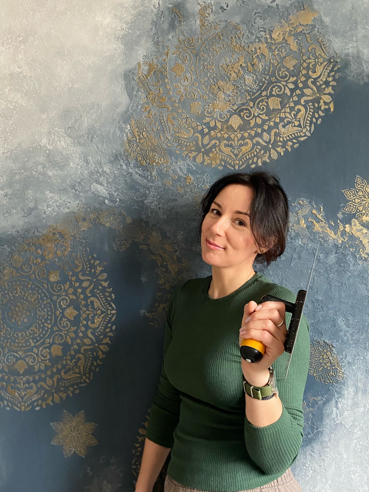 Dekoracyjna ściana z efektem ombre i złotymi ornamentami. Kobieta trzyma szpachelkę, prezentując efektowną technikę wykończenia wnętrz.