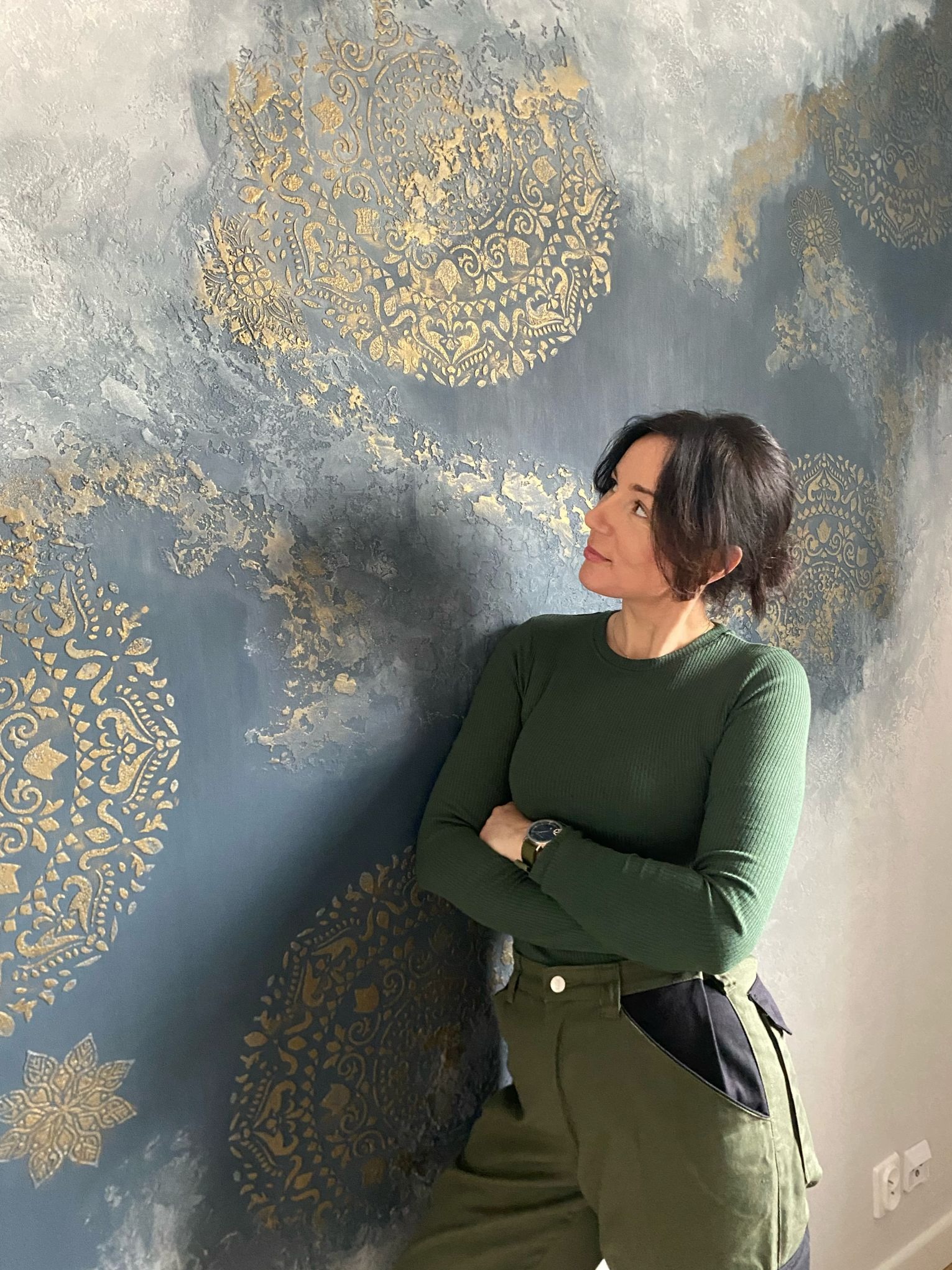 Dekoracyjna ściana z efektem ombre w odcieniach szarości i złota, zdobiona ornamentami. Kobieta opiera się o ścianę, prezentując efekt finalny.