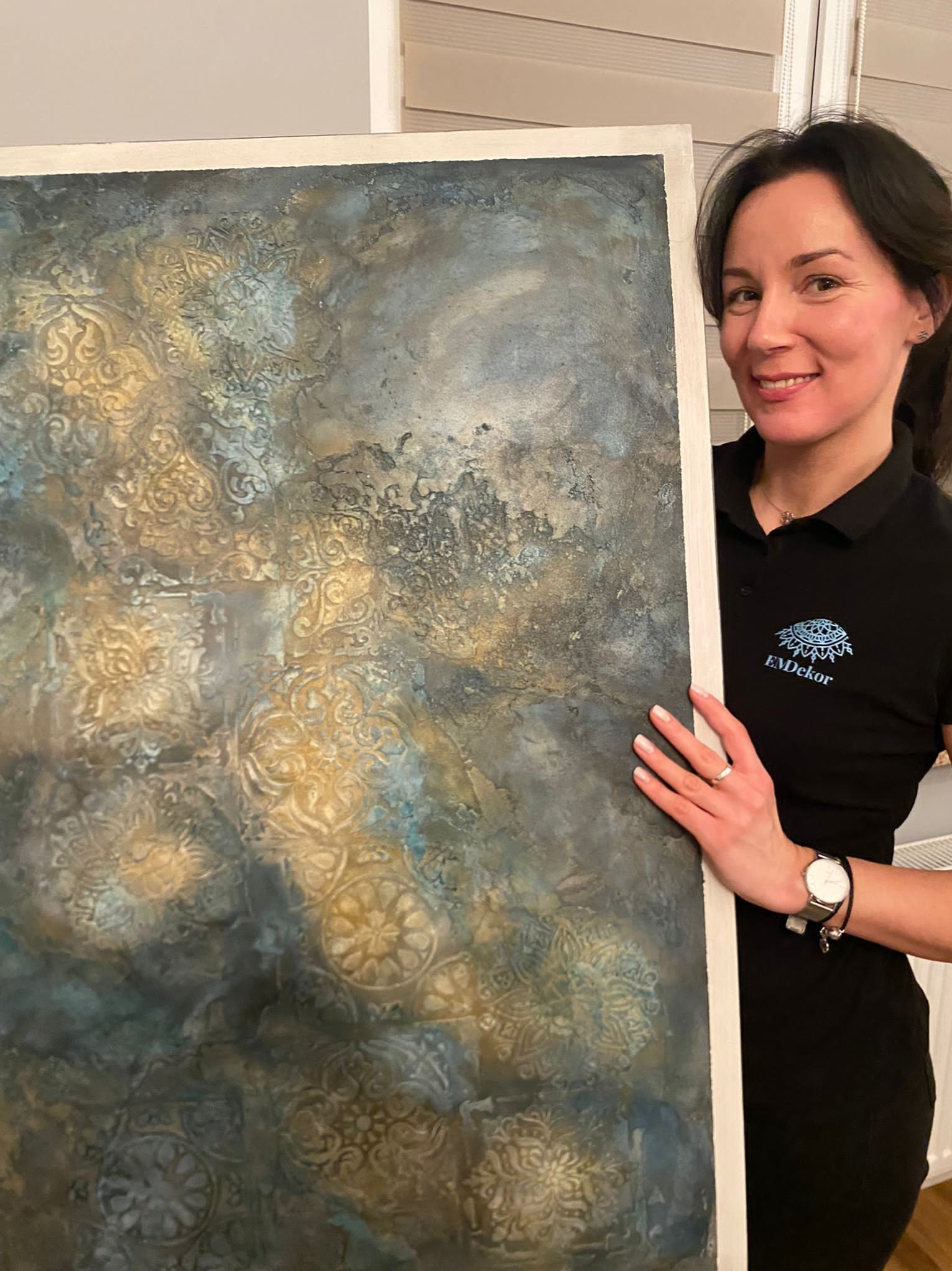 Dekoracyjna ściana z efektem tynku strukturalnego w odcieniach złota i błękitu, prezentowana przez uśmiechniętą kobietę w koszulce z logo firmy.