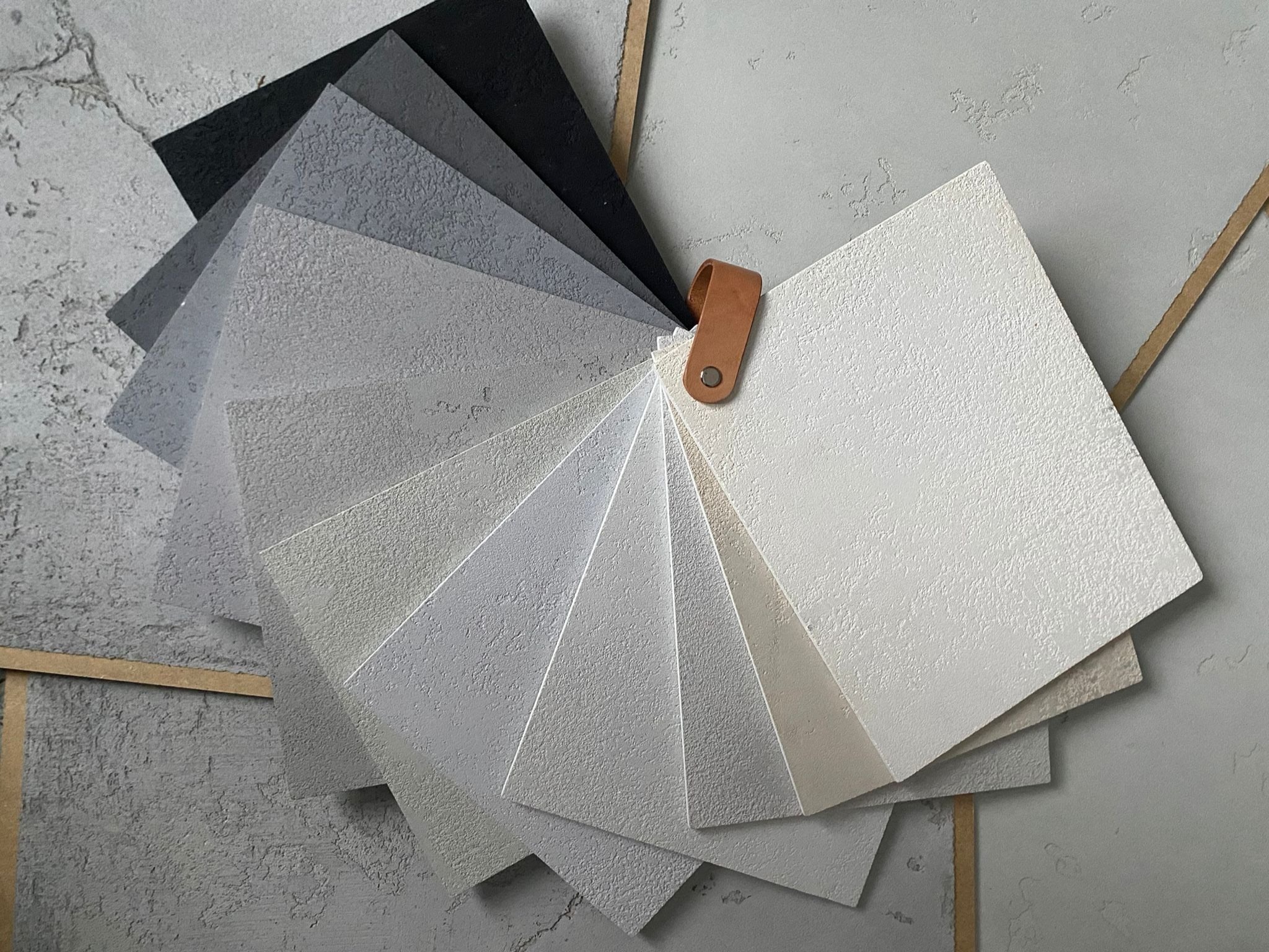 Paleta próbek tynków dekoracyjnych w odcieniach szarości i bieli, z wyraźną fakturą, połączona skórzanym uchwytem na tle ściany z efektem betonu.