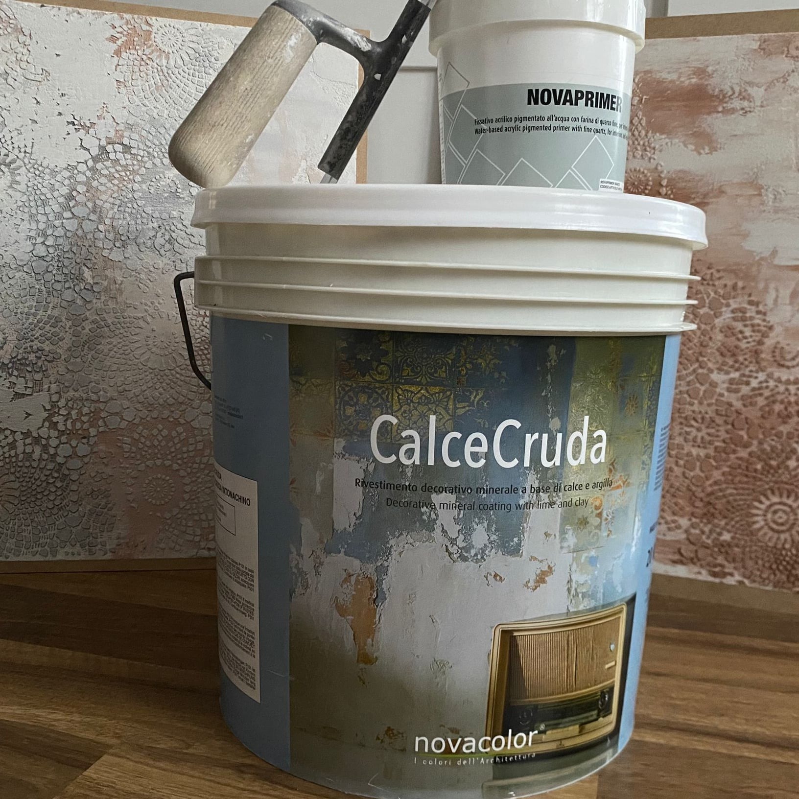 Wiadro z dekoracyjną farbą mineralną CalceCruda, obok puszka z gruntem Novaprimer i kielnia. W tle ściana z efektem dekoracyjnym, imitującym koronkę.