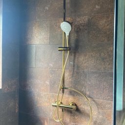 Usługi ogólnobudowlane - Nowoczesna kabina prysznicowa z deszczownicą w kolorze złotym, na tle ciemnych, imitujących kamień płytek. Minimalistyczny design, eleganckie wykończenie.