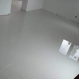 Usługi ogólnobudowlane - Błyszcząca, jasnoszara podłoga z płytek w pustym pomieszczeniu z białymi ścianami. Widoczne refleksy światła na gładkiej powierzchni. Minimalistyczny design.