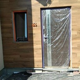 Usługi ogólnobudowlane - Elewacja budynku z oknem i drzwiami wejściowymi, wykończona płytkami imitującymi drewno. Drzwi zabezpieczone folią ochronną, widoczna instalacja elektryczna.