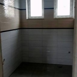 Usługi ogólnobudowlane - Wykończona łazienka z dwoma oknami, wyłożona kafelkami w odcieniach beżu i szarości z czarnym pasem, widok z wejścia.