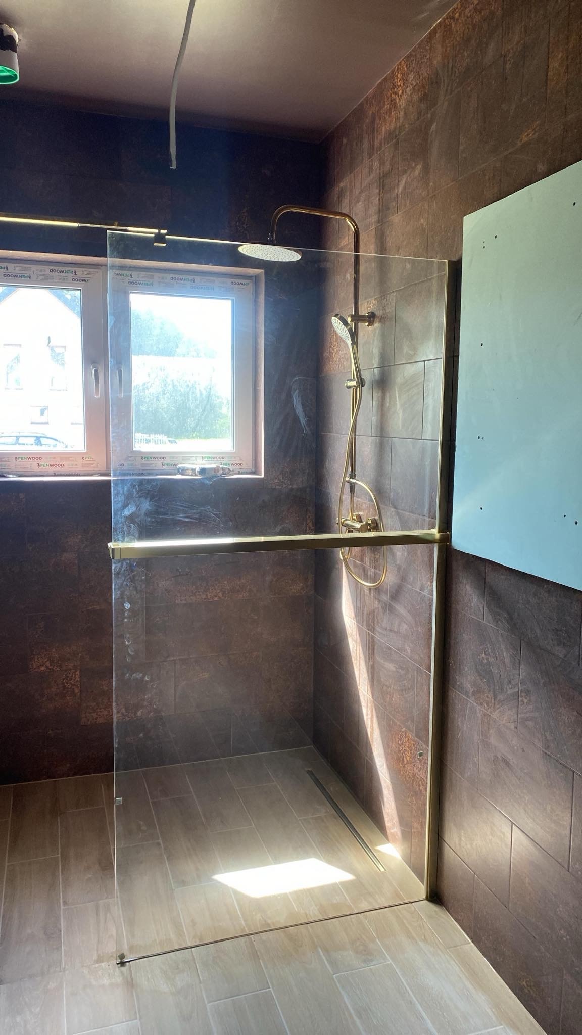 Nowoczesna kabina prysznicowa walk-in z złotymi elementami wykończeniowymi i brązowymi płytkami. Widoczne okno z widokiem na zewnątrz i fragment płyty gipsowej do wykończenia.