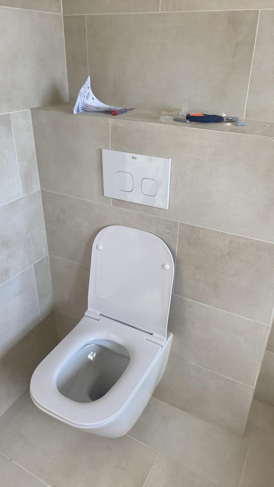 Zestaw podtynkowy WC z przyciskiem Roca, zamontowany na beżowych płytkach w łazience. Na półce instrukcja i narzędzia. Ukończona instalacja sanitarna.