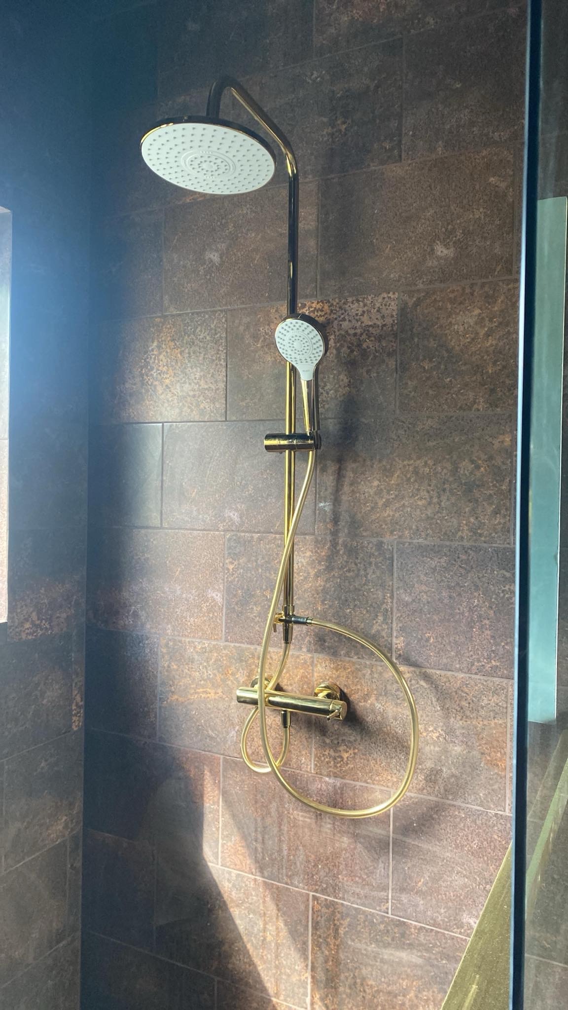 Nowoczesna kabina prysznicowa z deszczownicą w kolorze złotym, na tle ciemnych, imitujących kamień płytek. Minimalistyczny design, eleganckie wykończenie.