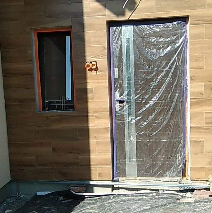 Elewacja budynku z oknem i drzwiami wejściowymi, wykończona płytkami imitującymi drewno. Drzwi zabezpieczone folią ochronną, widoczna instalacja elektryczna.