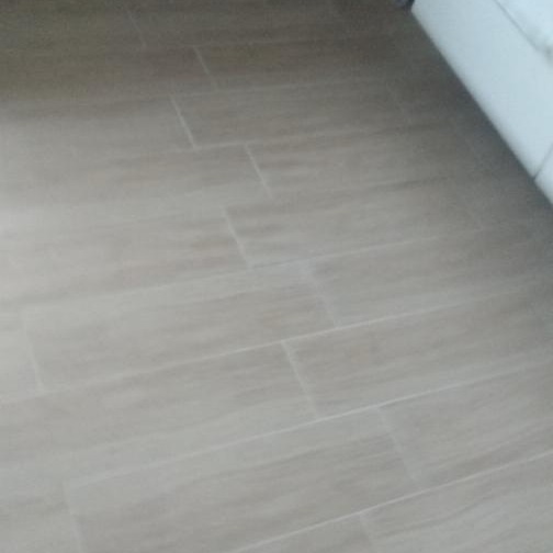 Ułożona podłoga z beżowych, prostokątnych płytek ceramicznych imitujących drewno, z widocznymi fugami i fragmentem białej listwy przypodłogowej w rogu.