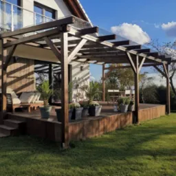 Drewniany taras przydomowy z zadaszeniem z przezroczystych płyt, otoczony zielonym trawnikiem, z meblami ogrodowymi i roślinami w donicach, widok na ogród.