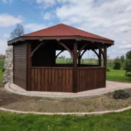 Ośmioboczna, ciemnobrązowa konstrukcja z czerwonym dachem, częściowo obłożona kamieniem, otoczona trawnikiem z kwitnącymi mniszkami, wykończona kostką brukową.