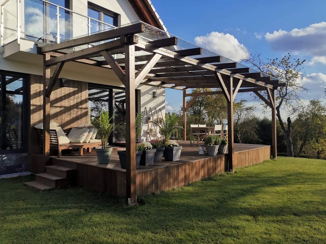 Drewniany taras przydomowy z zadaszeniem z przezroczystych płyt, otoczony zielonym trawnikiem, z meblami ogrodowymi i roślinami w donicach, widok na ogród.