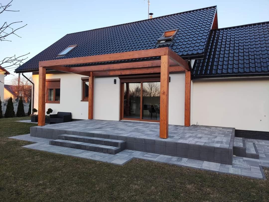 Nowoczesna pergola z drewna na podwyższonym tarasie z szarej kostki brukowej, przylegająca do białego domu z ciemnym dachem.