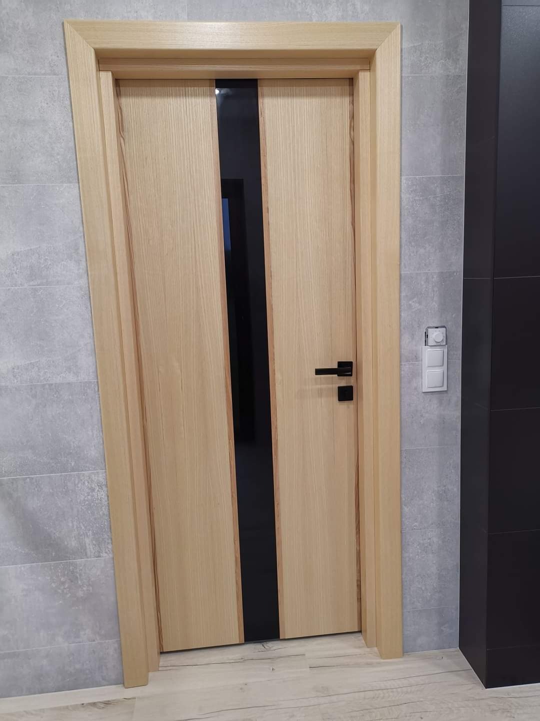Jasne, drewniane drzwi wewnętrzne z czarnym, pionowym pasem pośrodku, zamontowane w szarej ścianie z płytkami, widoczny czarny uchwyt i włącznik światła.