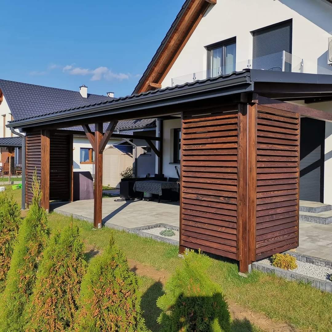 Zadaszona konstrukcja z drewna z lamelami, przylegająca do nowoczesnego domu z białymi ścianami i czarnym dachem, widoczna z perspektywy ogrodu z niskimi drzewkami iglastymi. Konstrukcja ma ciemny,...