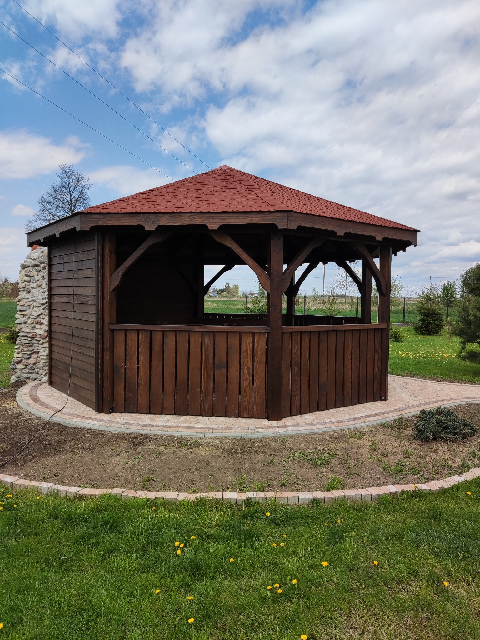 Ośmioboczna, ciemnobrązowa konstrukcja z czerwonym dachem, częściowo obłożona kamieniem, otoczona trawnikiem z kwitnącymi mniszkami, wykończona kostką brukową.