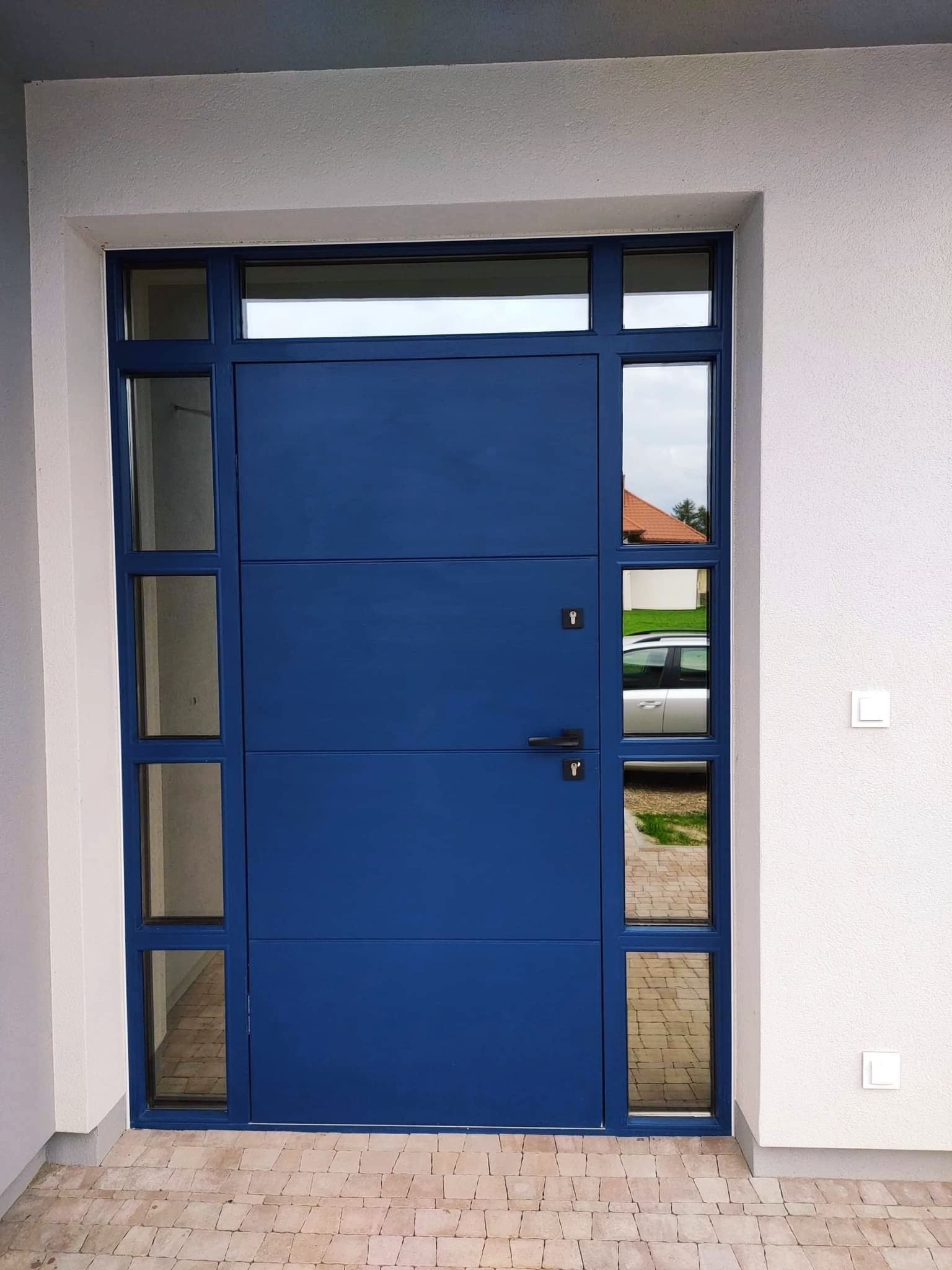 Nowoczesne, niebieskie drzwi wejściowe z bocznymi przeszkleniami, widoczne z zewnątrz, z kostką brukową u dołu i białym tynkiem na ścianie budynku.