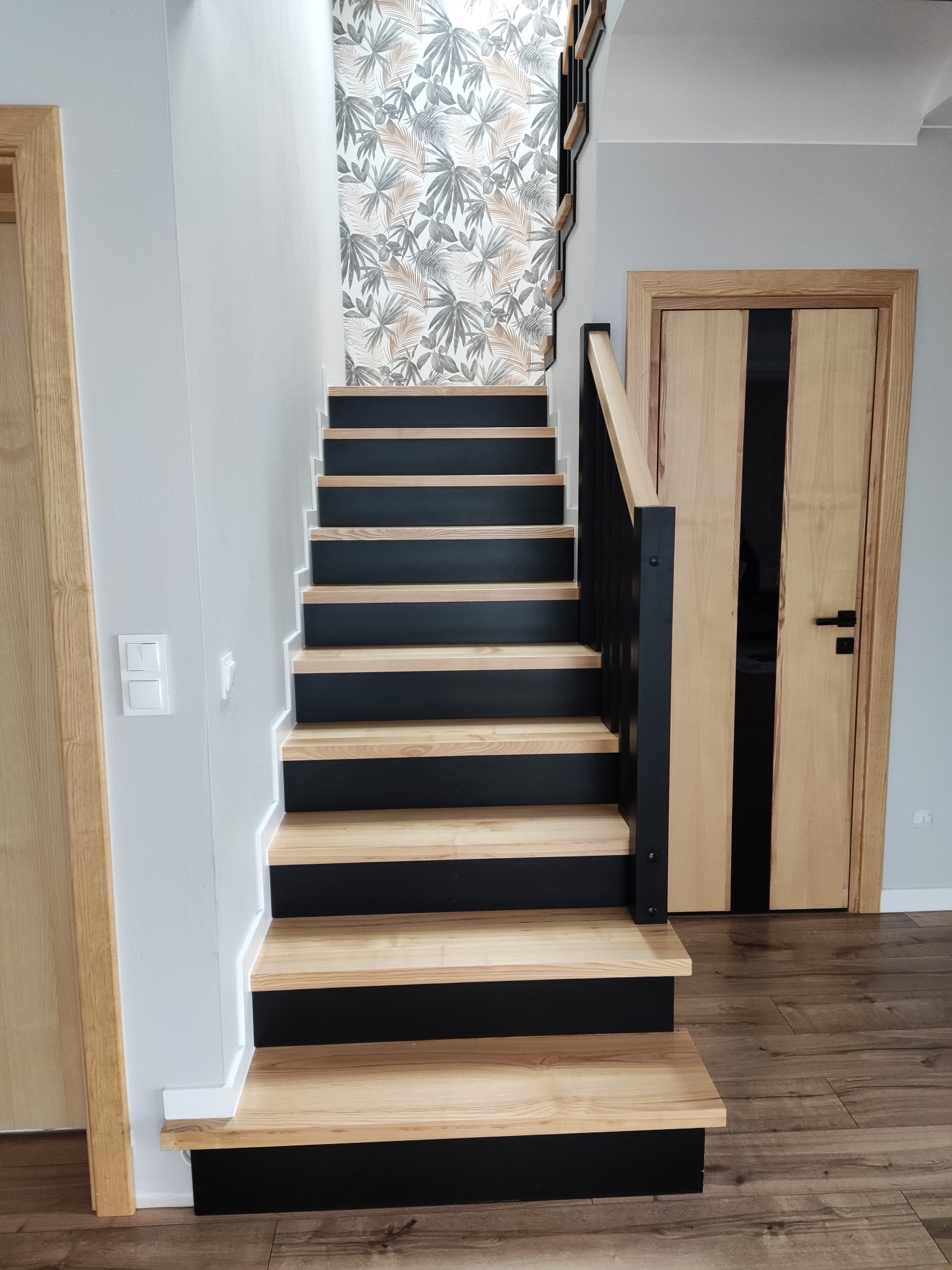 Schody drewniane z czarnymi podstopnicami i balustradą, widok od dołu, z tapetą w liście i drzwiami z jasnego drewna w tle.