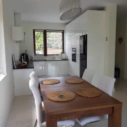 Nawymiar - Jasna kuchnia z białymi szafkami i czarnym blatem, drewniany stół z czterema plecionymi podkładkami i białymi krzesłami, okno z żaluzjami i ozdobny żyrandol.