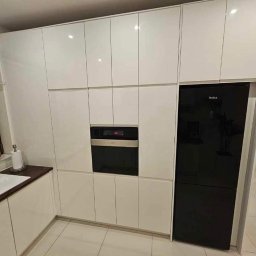 Nawymiar - Nowoczesna, biała zabudowa kuchenna z połyskiem, z czarnym piekarnikiem i lodówką Amica, widok na blat z dynią przy oknie.