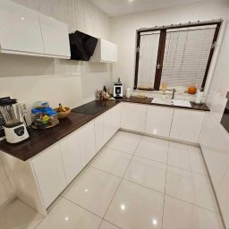 Nawymiar - Jasna kuchnia z białymi szafkami i ciemnym drewnianym blatem, widok na białe płytki podłogowe, okno z żaluzjami i dekoracjami świątecznymi, blender na blacie.