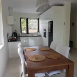 Jasna kuchnia z białymi, lakierowanymi frontami szafek, drewnianym stołem z czterema białymi krzesłami i okrągłymi podkładkami, oświetlona dekoracyjnym żyrandolem