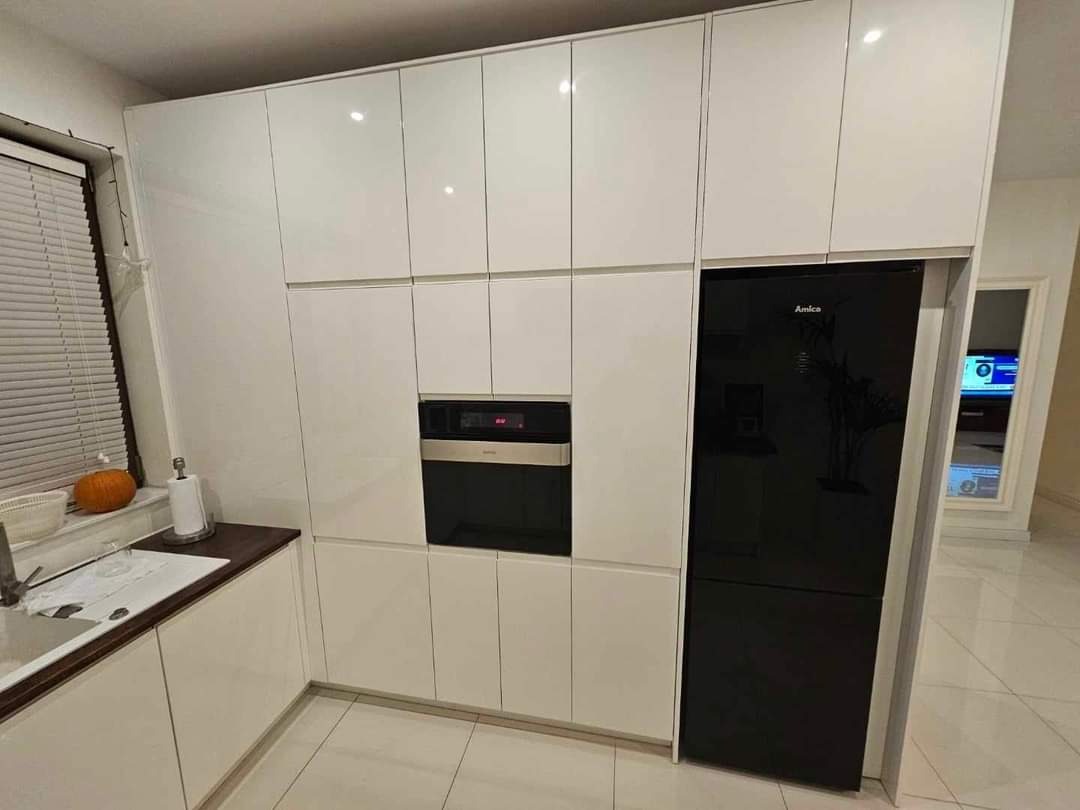 Nowoczesna, biała zabudowa kuchenna z połyskiem, z czarnym piekarnikiem i lodówką Amica, widok na blat z dynią przy oknie.