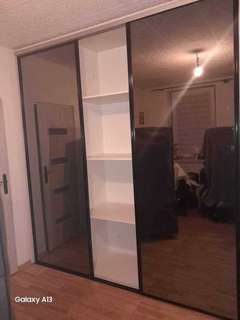 Szafa wnękowa z przesuwnymi drzwiami lustrzanymi i białymi półkami wewnętrznymi, widoczna na tle pokoju z drewnianą podłogą i drzwiami.