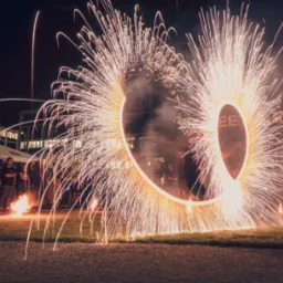 Nocny pokaz pirotechniczny z użyciem płonących kół, iskry rozświetlają publiczność na tle budynku z oknami.