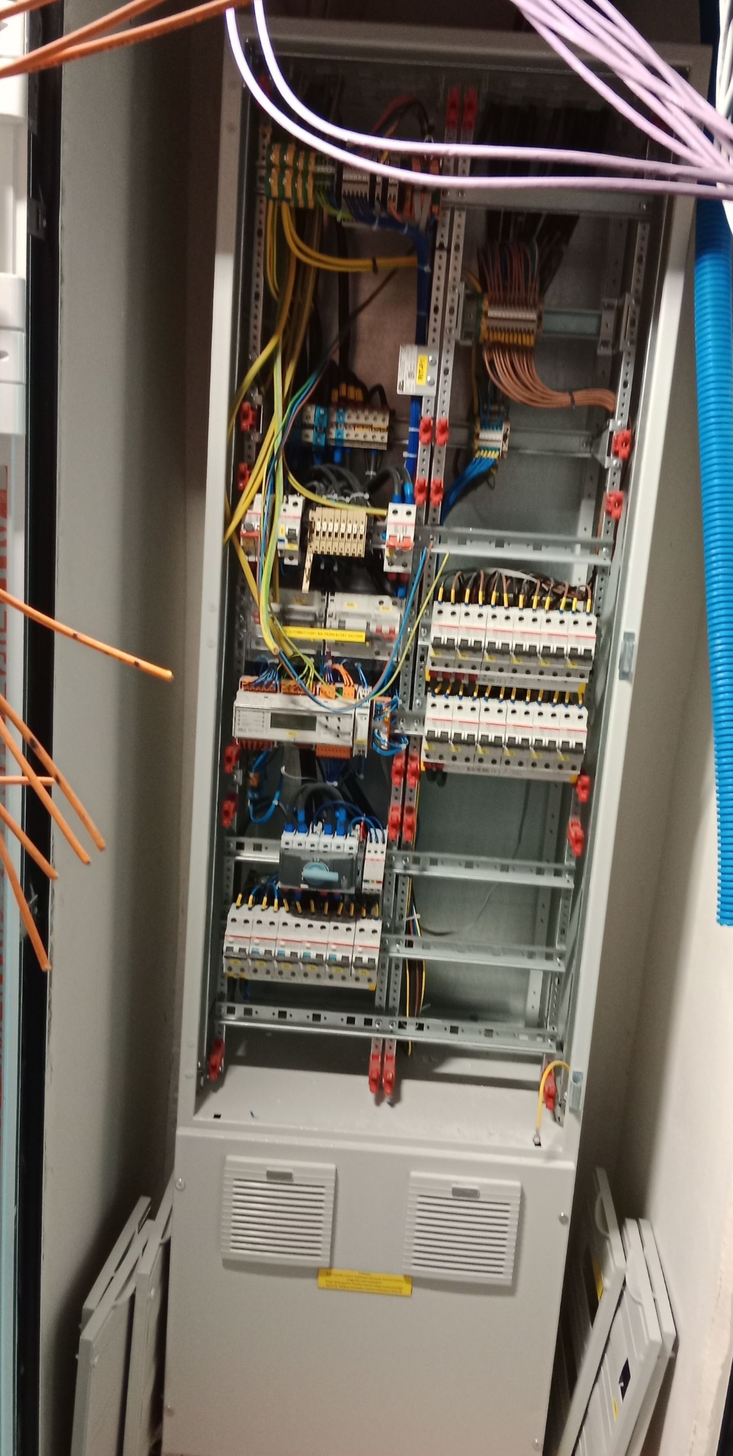 Otwarta szafa elektryczna z widocznymi bezpiecznikami, przewodami i listwami zaciskowymi, z pomarańczowymi kablami wystającymi z lewej strony i niebieską rurą karbowaną po prawej stronie.