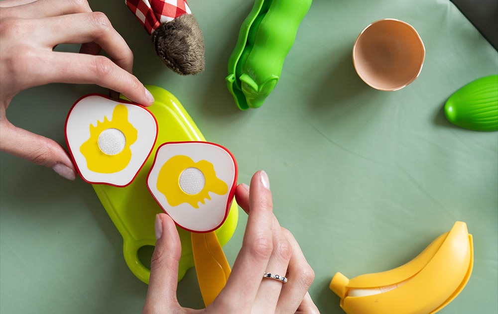 Dwie dłonie trzymają zabawki w kształcie jajek sadzonych na zielonej desce do krojenia; w tle zabawki przedstawiające warzywa i owoce.