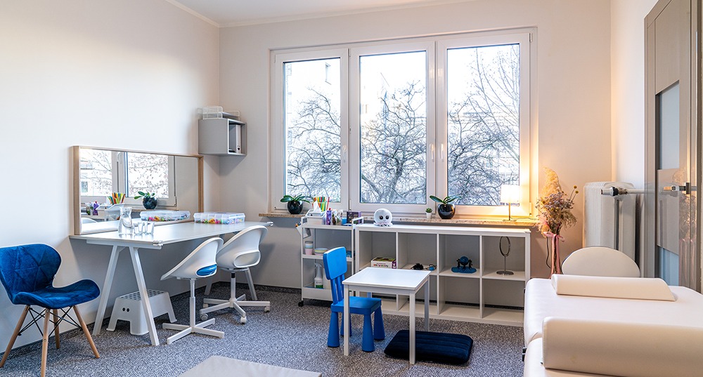 Jasny gabinet logopedyczny z biurkiem, lustrem, pomocami dydaktycznymi i oknem z widokiem na drzewa bez liści. Dodatkowe małe biurko i krzesło dla dziecka.