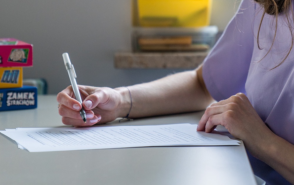 Osoba pisze na kartce papieru srebrnym długopisem; w tle widoczne gry planszowe i półka z pudełkami.