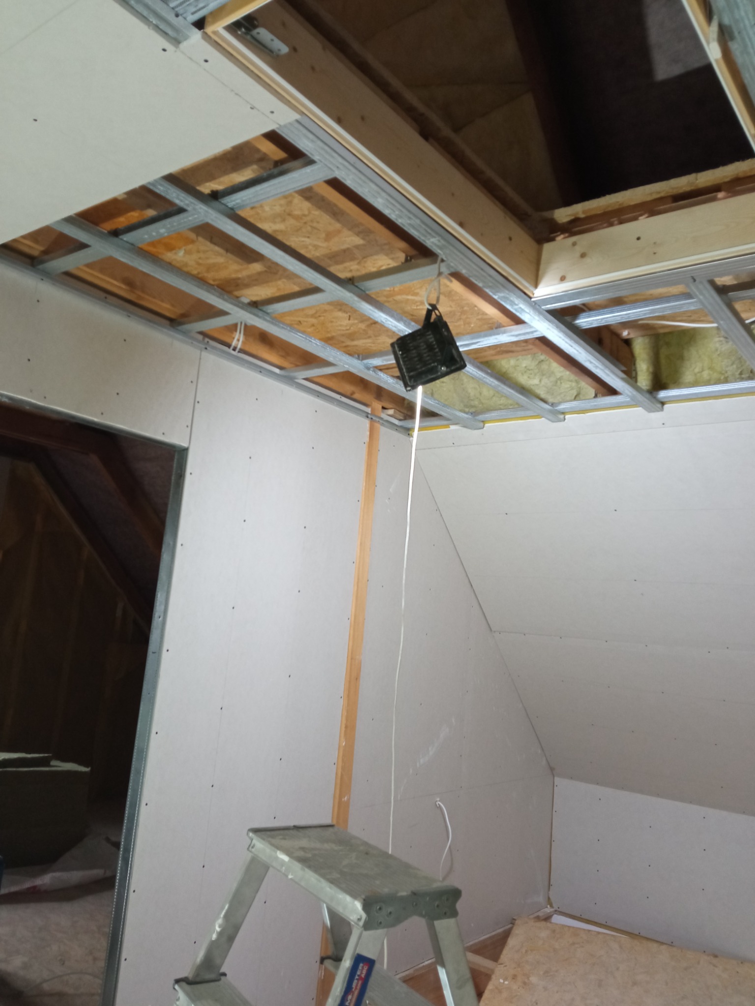 Montaż ścian działowych na poddaszu: widoczna metalowa konstrukcja sufitu, płyty w trakcie mocowania, drabina aluminiowa w rogu pomieszczenia, tymczasowe oświetlenie robocze zwisające z konstrukcji.