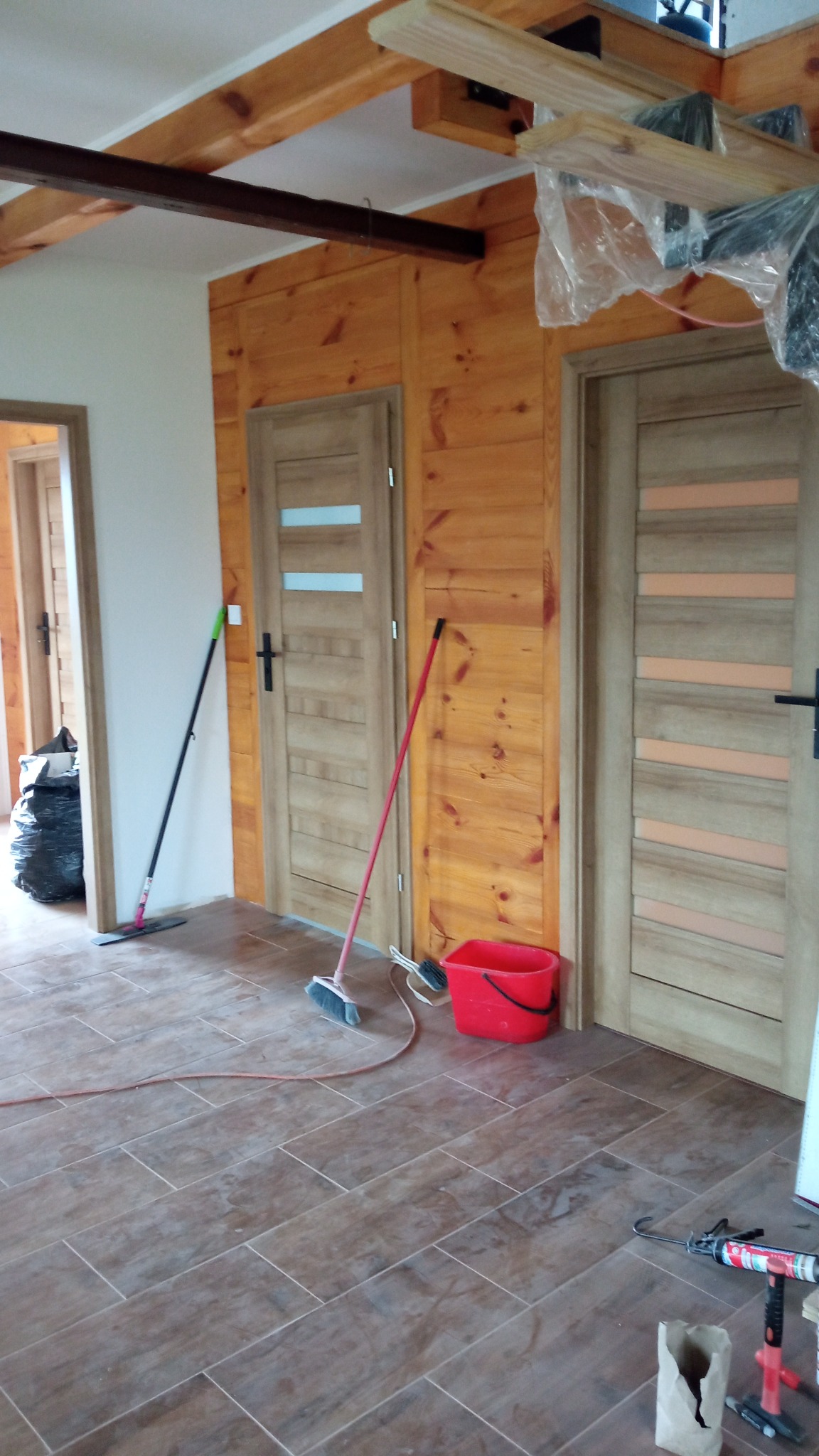 Wnętrze z drewnianą boazerią i dwiema parami drzwi z jasnego drewna, widoczne narzędzia i akcesoria malarskie sugerują prace wykończeniowe, z podłogą wyłożoną brązowymi płytkami imitującymi drewno.