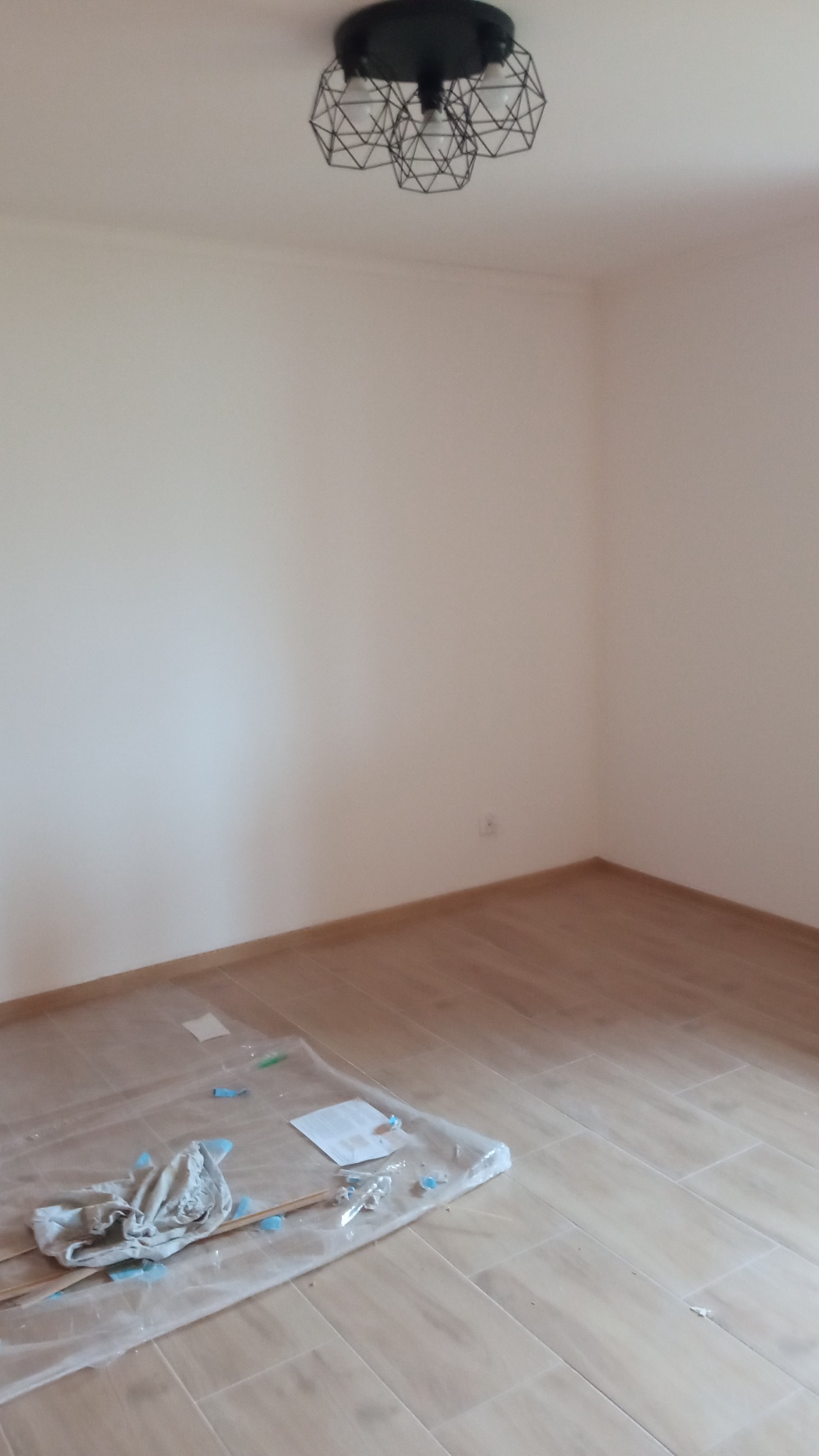 Pusta ściana po świeżym pomalowaniu w pokoju z płytkami imitującymi drewno na podłodze, zabezpieczonymi folią malarską z resztkami farby i narzędziami, nowoczesna lampa sufitowa w stylu industrialnym.
