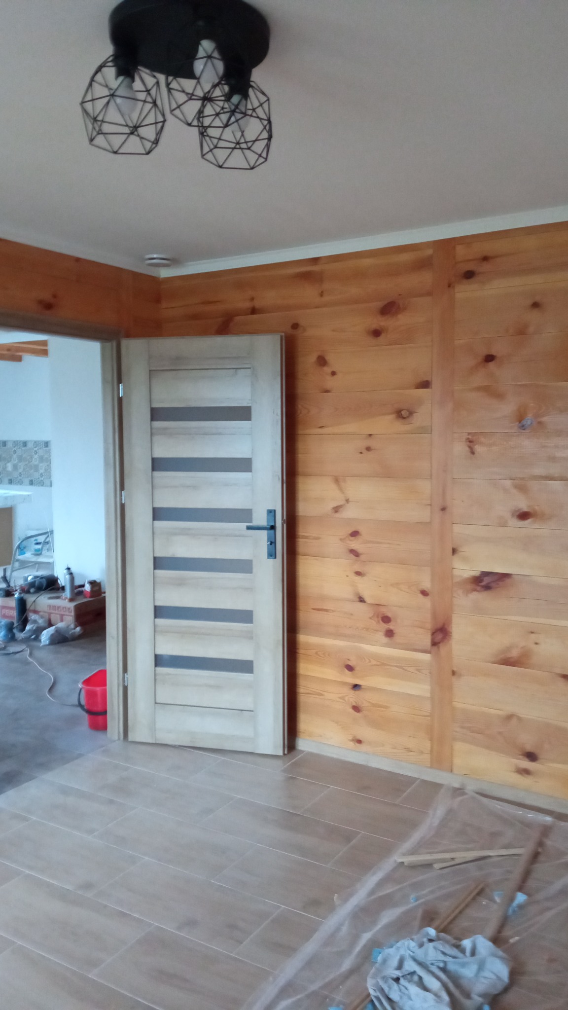 Wykończone wnętrze w Lublinie z drewnianą boazerią, nowoczesną lampą wiszącą i nową podłogą z płytek ceramicznych, widoczna otwarta przestrzeń z elementami kuchni w tle.