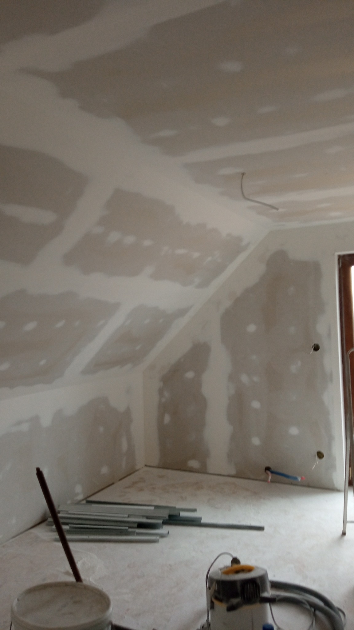 Poddasze w trakcie prac wykończeniowych: częściowo wyszpachlowane ściany i sufit, widoczne profile, wiadro i odkurzacz przemysłowy.