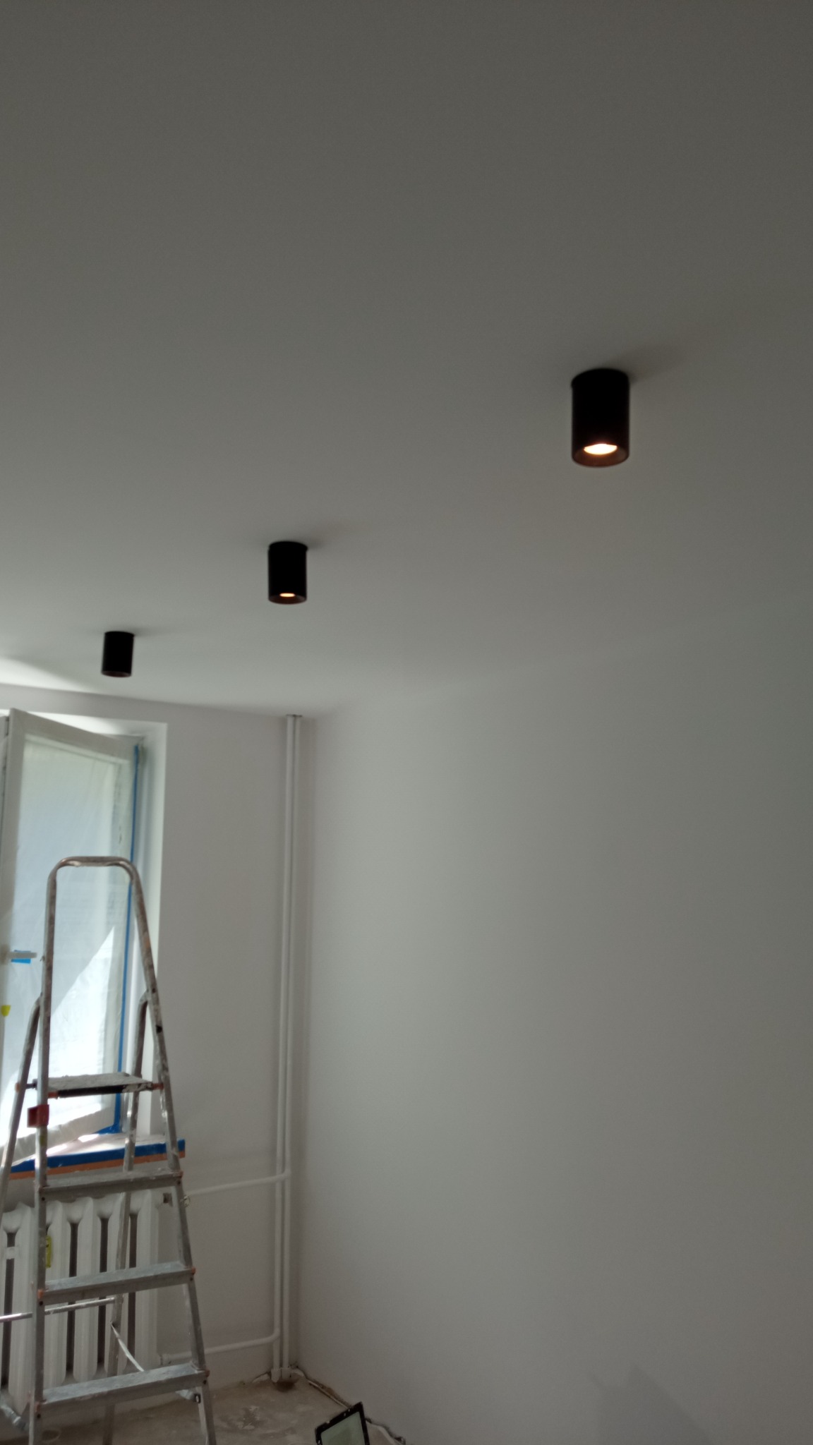 Wnętrze po remoncie: Świeżo pomalowane białe ściany z trzema czarnymi, cylindrycznymi lampami sufitowymi, drabina oparta o okno z widocznym grzejnikiem.
