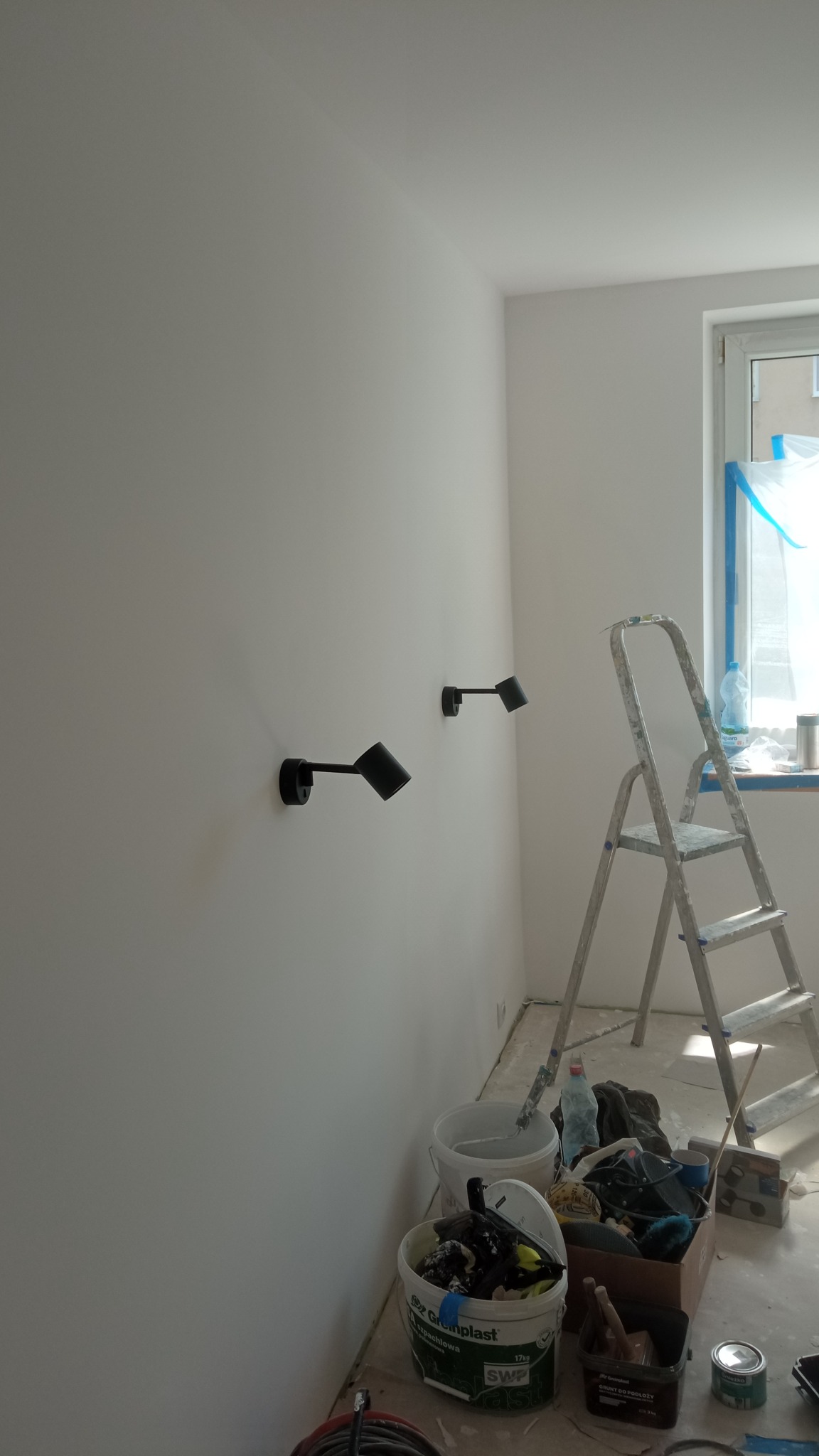 Świeżo pomalowana na biało ściana z zamontowanymi dwoma czarnymi kinkietami, widoczne ślady po pracach malarskich, drabina aluminiowa, wiadro z farbą i akcesoria malarskie przy ścianie.