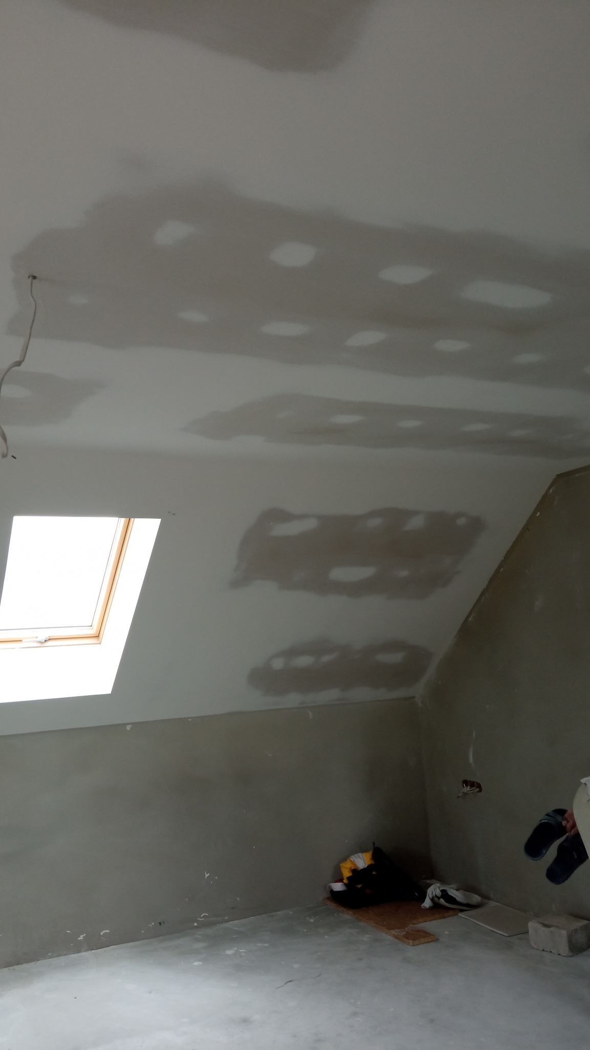 Poddasze w trakcie wykańczania: sufit ze śladami po łataniu, ściany przygotowane pod malowanie, widoczne okno dachowe i porozrzucane narzędzia w rogu pokoju.
