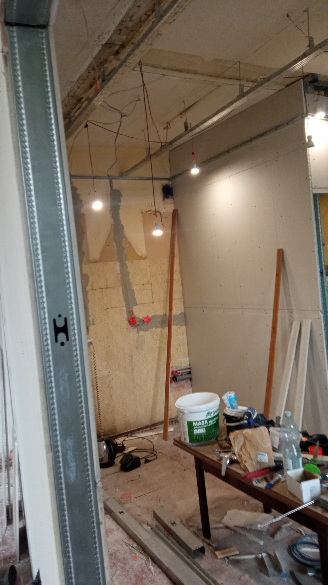 Wnętrze w trakcie remontu: widoczne profile metalowe, częściowo położona płyta na ścianie, odsłonięta instalacja elektryczna, narzędzia i materiały budowlane na stole, surowe ściany w tle.