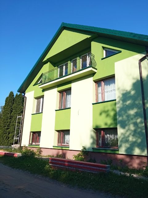 Dwupiętrowy dom z balkonem, pomalowany w zielono-kremowe pasy, z aluminiową drabiną opartą o ścianę.