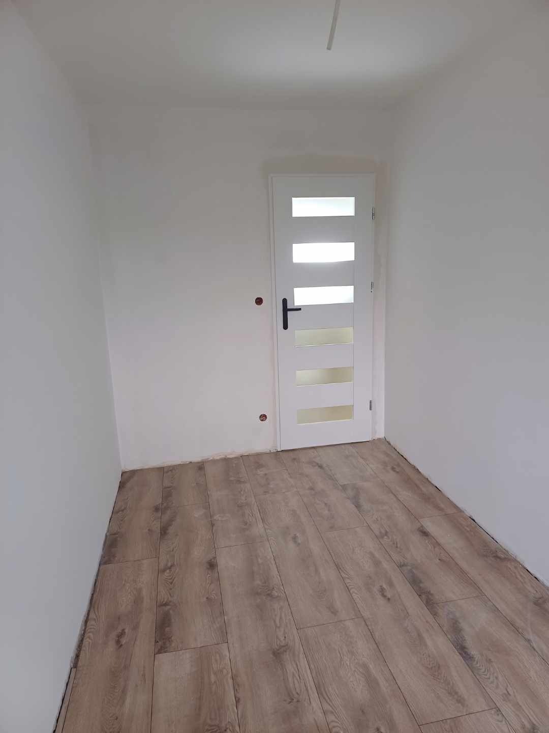 Wnętrze pokoju po remoncie: ułożone panele podłogowe w odcieniach brązu, białe ściany i białe drzwi z czarną klamką i przeszkleniami. Widoczne niedoskonałości przy listwach przypodłogowych.