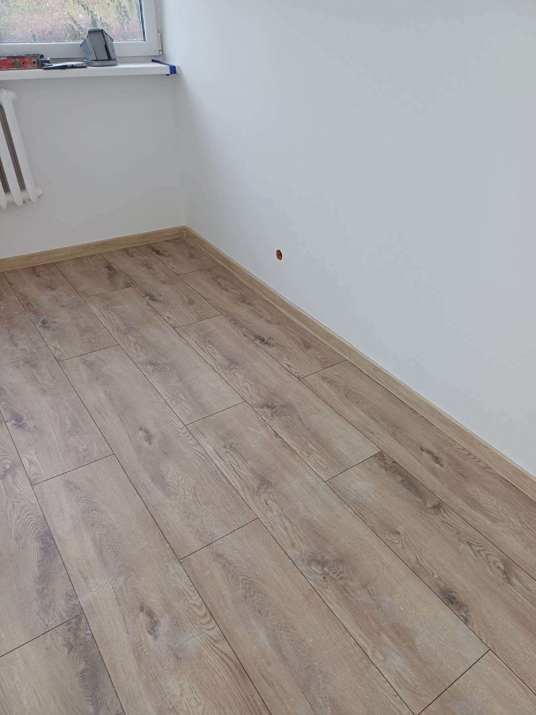 Ułożone panele podłogowe w jasnym odcieniu drewna w pomieszczeniu w Bydgoszczy, widoczna ściana z otworem na gniazdko.