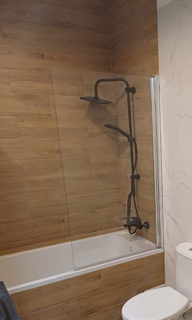 Wykończona łazienka z wanną obudowaną płytkami imitującymi drewno, czarna armatura natryskowa i szklany parawan nawannowy.